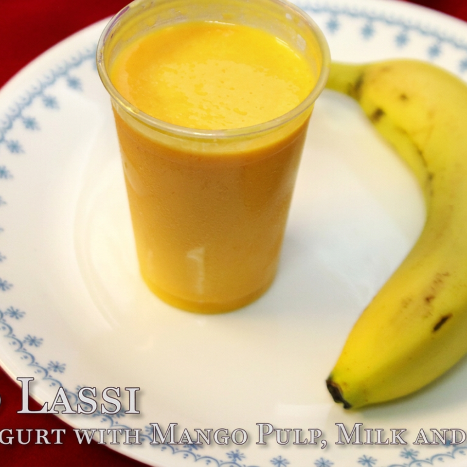 Glass of mango lassi beside a ripe banana on a blue-patterned plate against a red tablecloth.